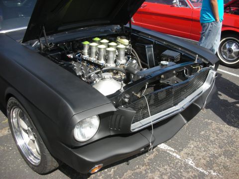 2004 Ford Crown Victoria - A black 1965 Ford Mustang's engine bay is open, revealing its custom engine, with a red 1965 Ford Mustang visible behind it.