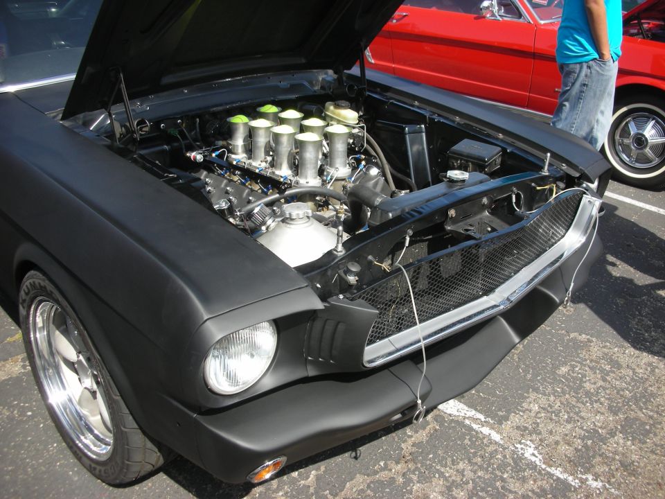 Black 1965 Ford Mustang engine bay with chrome intake stacks and a red 1965 Ford Mustang in the background