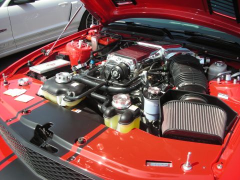 2004 Ford Crown Victoria - Engine bay of a red car with a prominent supercharger and black racing stripes, with a white car visible in the background.