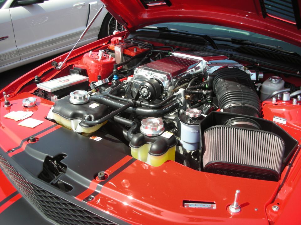 Red engine bay with supercharger and black racing stripes. White car in background