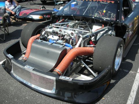 2004 Ford Crown Victoria - A racing car with its hood open reveals a complex engine and orange hoses, parked at an outdoor event.