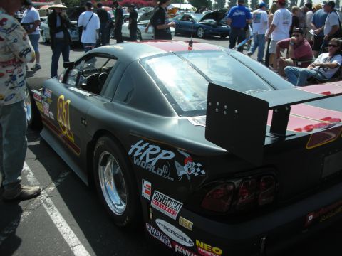 2004 Ford Crown Victoria - A dark-colored race car with sponsor decals and a prominent rear wing is parked at an outdoor event.