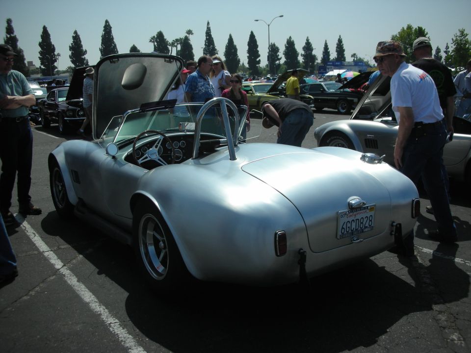 Silver Shelby Cobra replica with hood open, people looking at it and other cars in background