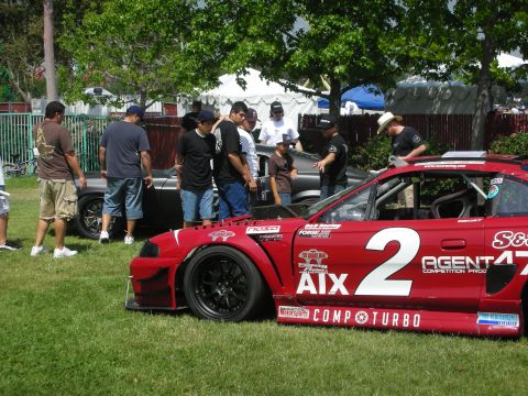 2004 Ford Crown Victoria - A red race car with racing decals and a large number 2 is parked on grass with spectators around.