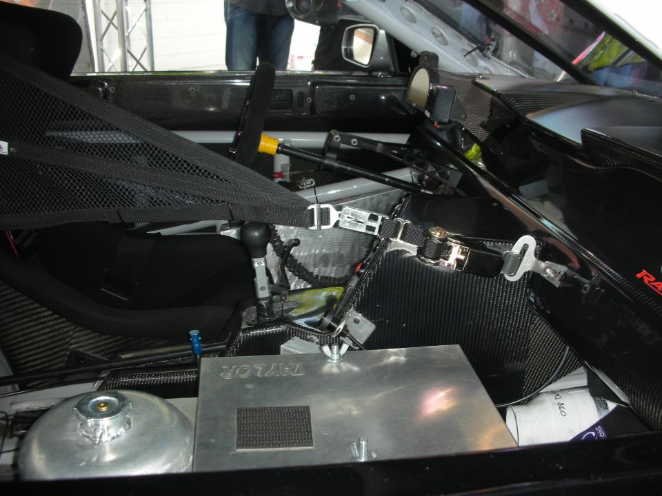 Interior of a race car showing a racing seat, roll cage, shifter, and a metal box labeled TAYLOR