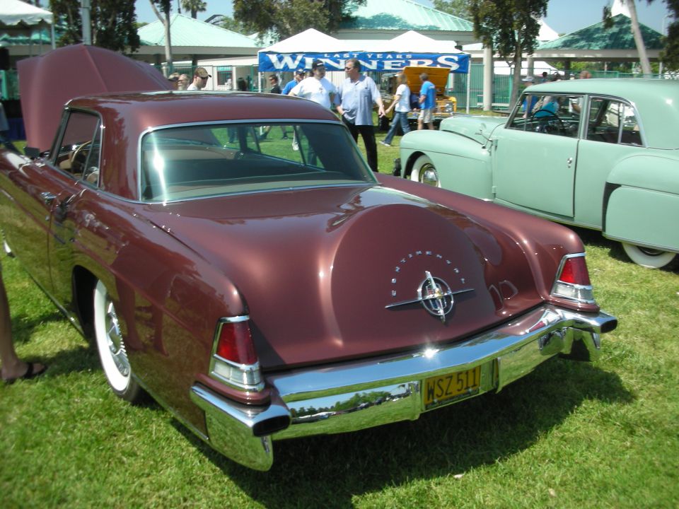 Rear view of a maroon 1956 Lincoln coupe with its hood up, parked on grass next to a light green sedan