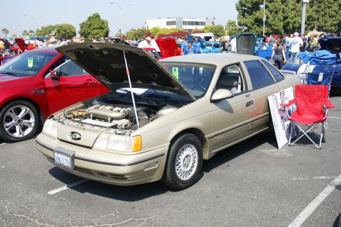 1992 Ford Taurus - A tan 1992 Ford Taurus sedan is displayed with its hood open at a car show, next to a red folding chair.