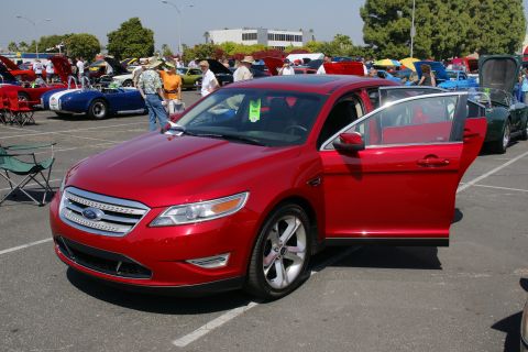 1992 Ford Taurus - A red sedan is displayed with its doors open at a car show, surrounded by other vehicles and attendees.