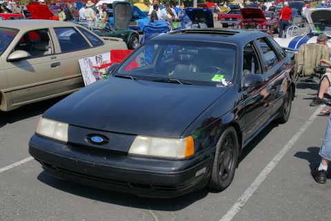 1992 Ford Taurus - A black 1992 Ford Taurus sedan is displayed at a car show, with other vehicles and people visible in the background.