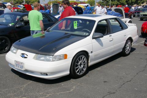 1992 Ford Taurus - White 1992 Ford Taurus sedan with aftermarket black hood and racing wheels at a car show.