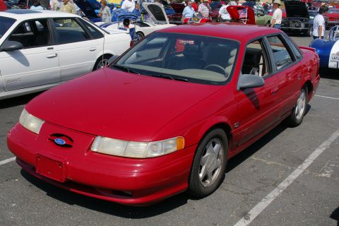 1992 Ford Taurus - A red sedan is parked in a lot, with other vehicles and attendees around it.