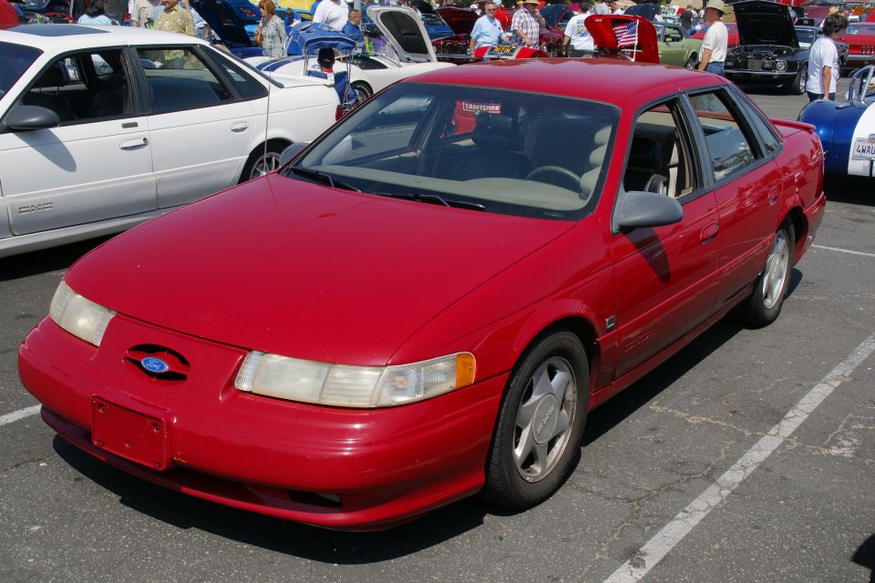 Red sedan parked at an outdoor car show. Other cars and people are visible in the background