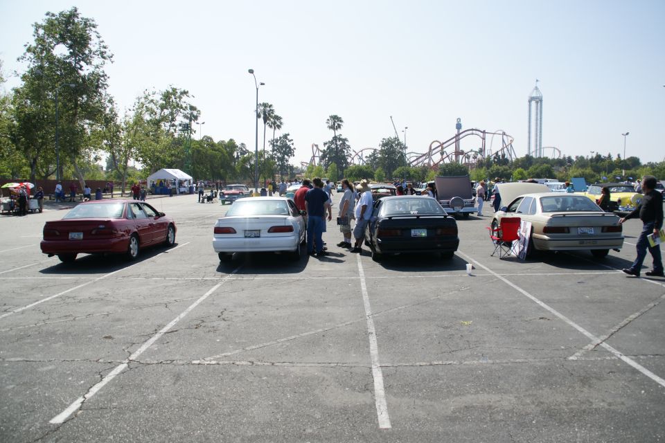 Several cars parked in a lot with people milling around. Roller coaster in background