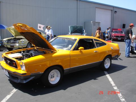 1978 Ford Mustang II - Yellow coupe with hood open, parked outdoors with other vehicles and people.