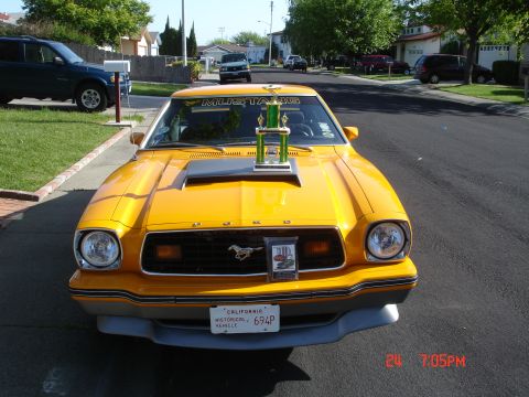 1978 Ford Mustang II - A yellow 1978 Ford Mustang II is parked on a residential street with a trophy displayed on its hood.