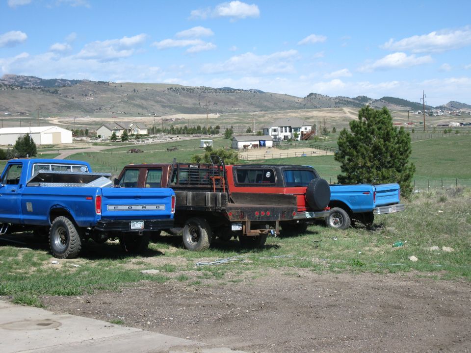 Three Ford trucks and a Bronco parked in a field with houses and hills in the background