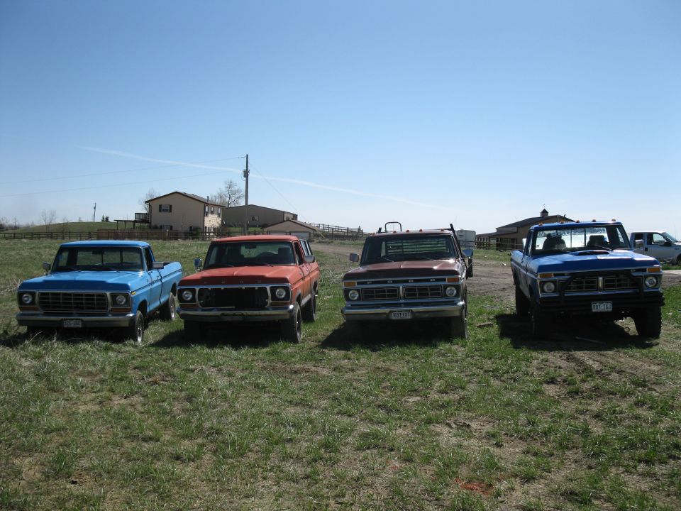 Four trucks and an orange SUV parked in a field. The blue truck on the far left looks like a 1978 Ford Bronco