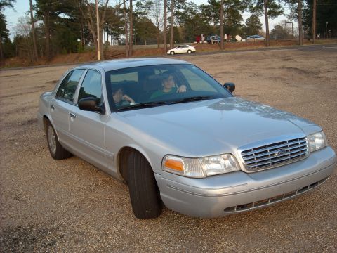 2003 Ford Crown Victoria - A silver 2003 Ford Crown Victoria with two flat tires is parked on a gravel lot, with two people visible inside.