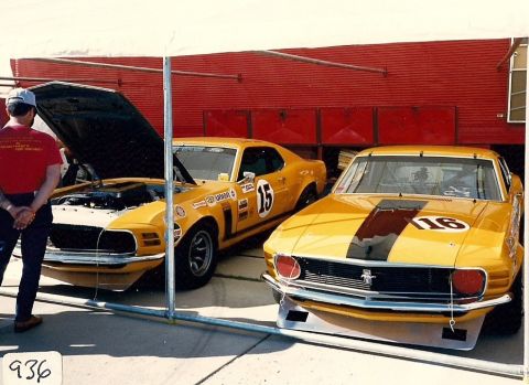 1973 Ford Mustang - Two yellow racing Mustangs are parked under a canopy, one with its hood open, revealing the engine.