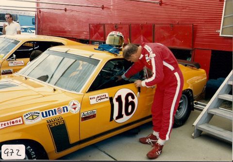 1973 Ford Mustang - A race car driver in a red suit attends to a yellow race car with 'Boss 302' decals and the number 16.