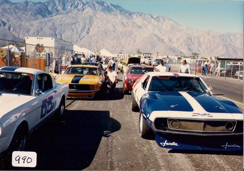 1973 Ford Mustang - A blue and white AMC Javelin race car is in the foreground on a track, with other cars and spectators in the background.