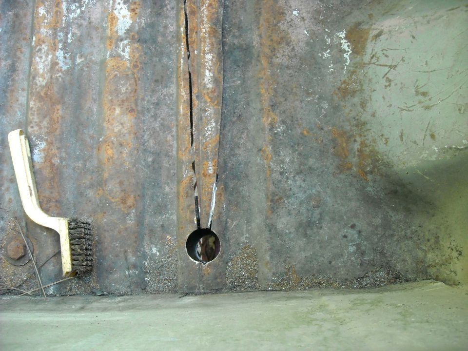 Close-up of a rusty metal panel with a crack and a hole, next to a cleaning brush