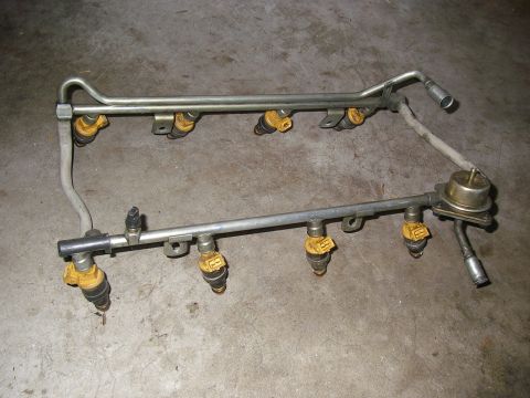 1990 Ford Bronco - A dual fuel rail assembly with eight yellow-topped fuel injectors and a round fuel pressure regulator, laid on a concrete floor. Metal supp…