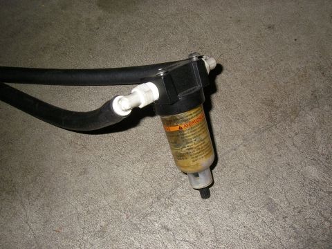 1990 Ford Bronco - Removed oil separator / catch can assembly lying on a concrete floor, its clear canister partly full of yellow-brown oily residue and weari…