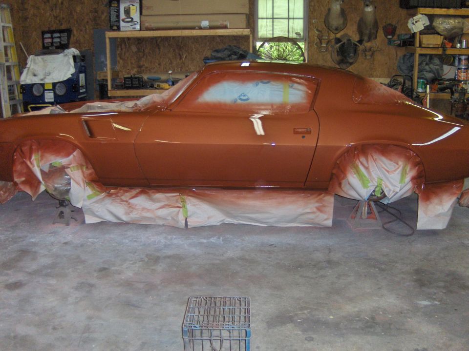 Orange coupe with wheels covered in paper, undergoing spray painting in a garage