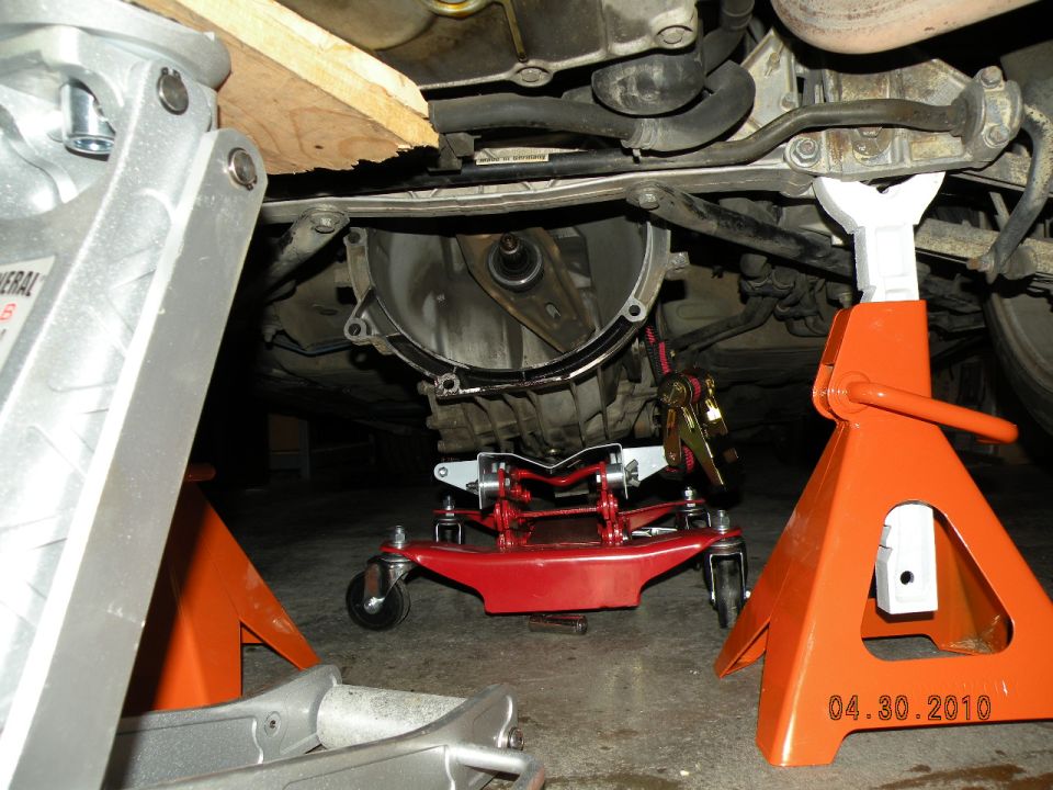 Car transmission supported by a red transmission jack and orange jack stands, with a wooden plank overhead