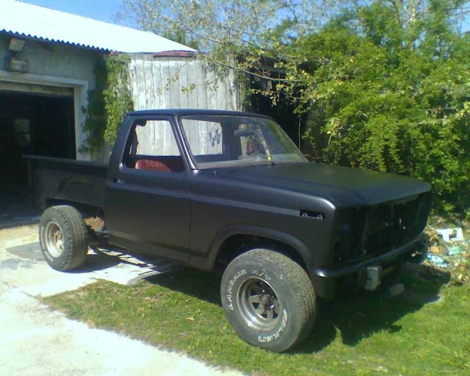 Black 1980 Ford F-100 pickup truck, missing grille, parked outside a garage