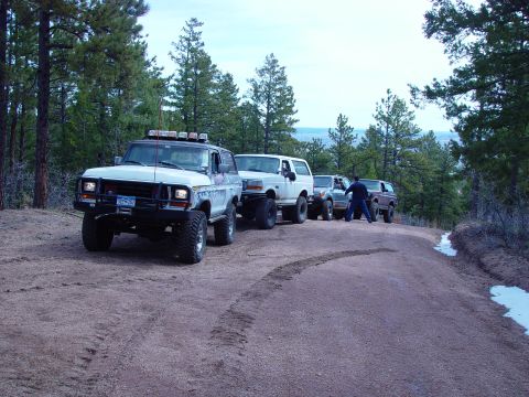 1993 Ford Bronco - A convoy of modified SUVs, including a white Ford Bronco, navigates a wooded trail.
