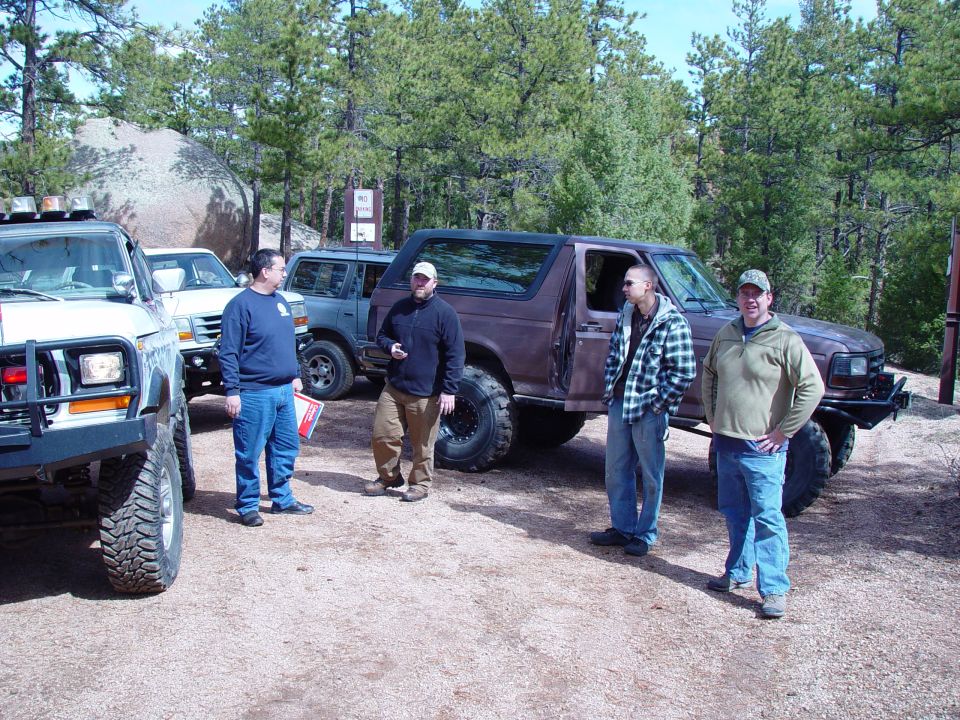 1992 Ford Bronco and other off-road vehicles parked with people standing around them in a wooded area
