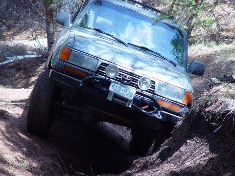 1993 Ford Bronco - A silver Toyota Land Cruiser navigates a challenging off-road trail, with its front-left wheel deep in a rut.