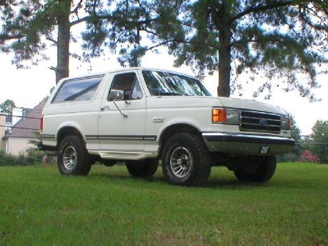 1989 Ford Bronco - A white Ford Bronco is shown from the front-right angle, parked on a grassy lawn with trees in the background.