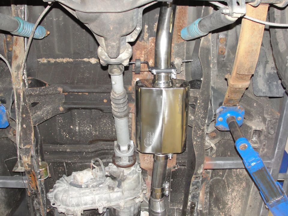 Undercarriage view of a shiny aftermarket exhaust muffler and pipes, with leaf springs and driveshaft visible