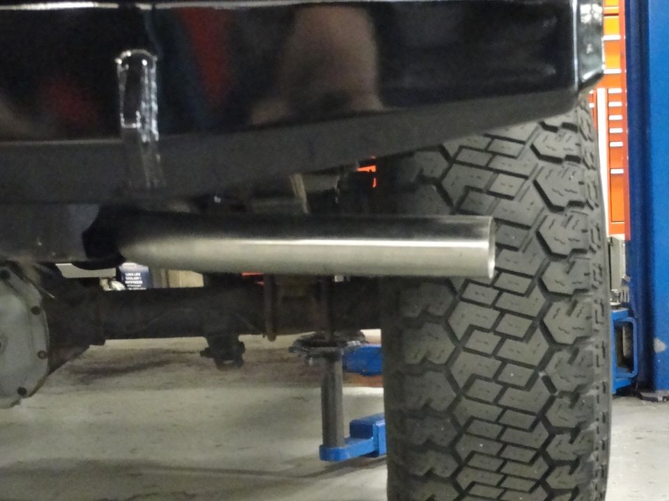 Close-up of a silver exhaust pipe extending from a black vehicle, with a large tire and suspension visible