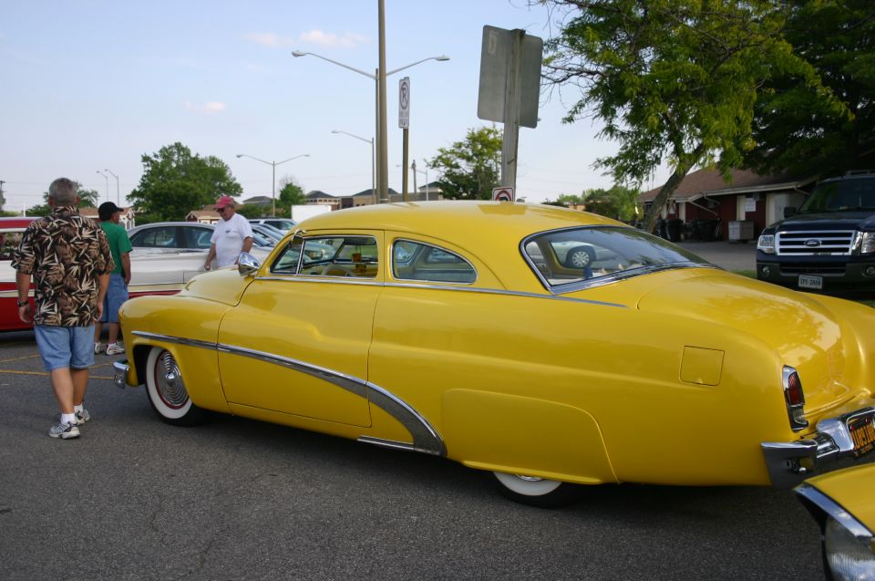 Yellow 1950s coupe with chrome side trim, seen from the rear three-quarter view