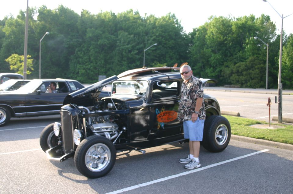 Guy next to a black hot rod with surfboards on top. Another car is parked to the left
