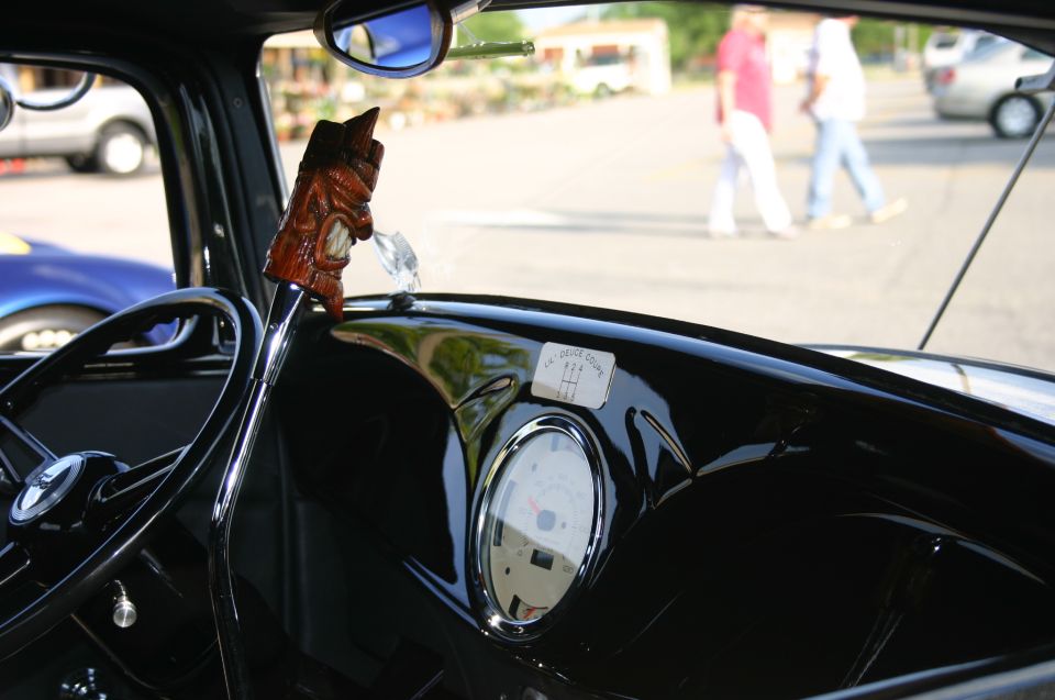 Dashboard view of a classic car with a tiki shift knob and a gauge cluster. People walk by outside