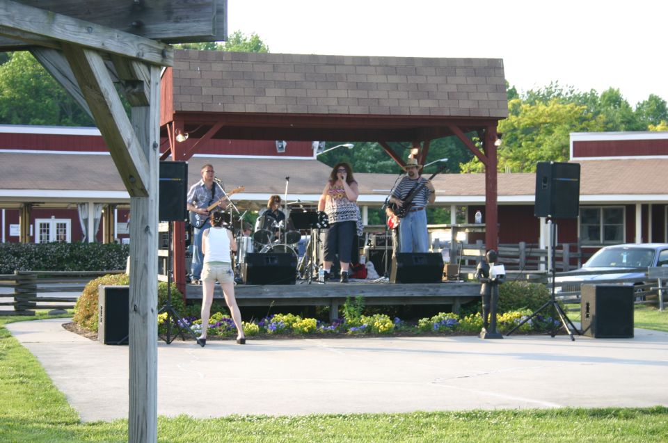 Band plays on a covered stage with a singer and guitarists. A car is parked to the right