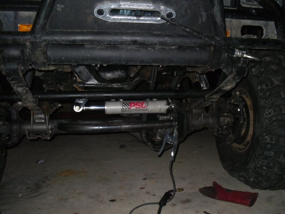 Close-up of a PSC steering stabilizer mounted to a vehicle's front axle with large tires and suspension components visible