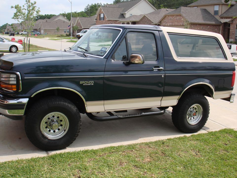Dark green and tan two-tone Ford Bronco with lift and oversized tires on a suburban driveway, side view