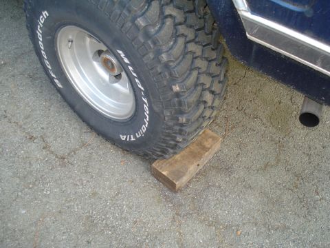 1984 Ford Bronco - Close-up of a tire on a blue vehicle, supported by a wooden block, with an exhaust pipe visible.