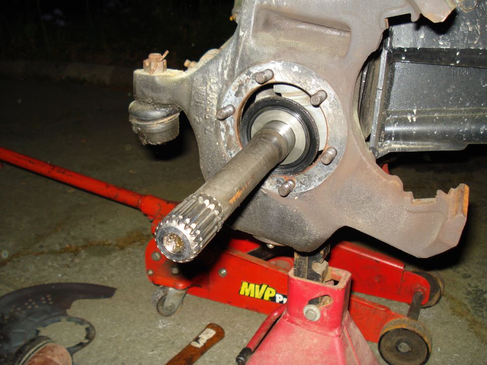 Close-up of a vehicle's brake assembly with a splined axle shaft extended. A red hydraulic jack and jack stand support the assembly