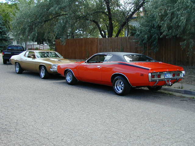 Orange 1971 Dodge Charger and gold 1974 Plymouth Satellite parked on street with black pickup truck behind them