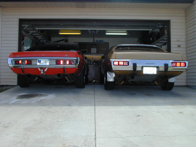 Two muscle cars, a red one and a gold one, parked in front of an open garage