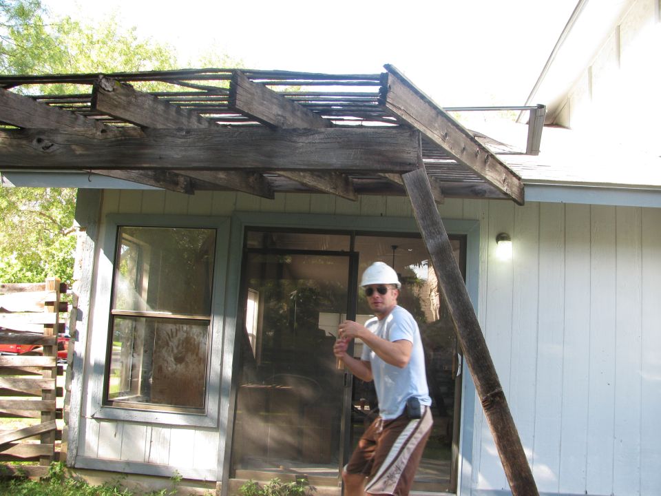 Man wearing a white hard hat and sunglasses stands near a damaged wooden pergola attached to a house