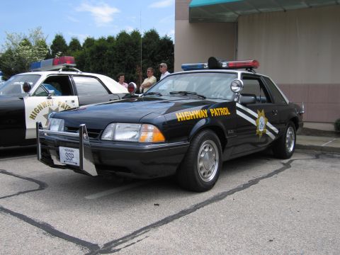 1991 Ford Crown Victoria - A black Ford Mustang SSP police car is parked on asphalt, with another police car visible to its left.