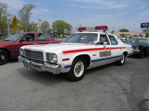 1991 Ford LTD Crown Victoria - white 1970s Plymouth full-size STATE POLICE sedan with red-and-blue stripes, a gold star and roof light bar parked at a curb.